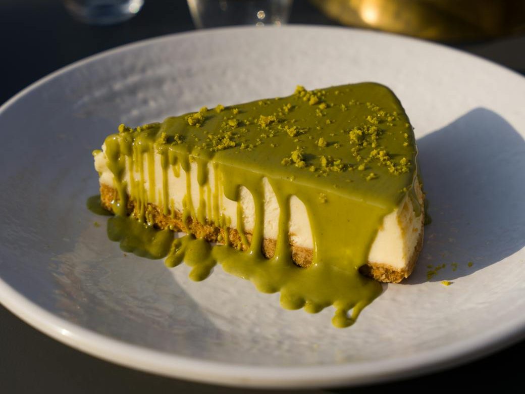 Potongan cheesecake matcha di atas piring putih, dengan saus matcha yang mengalir di permukaan dan taburan pistachio di atasnya.